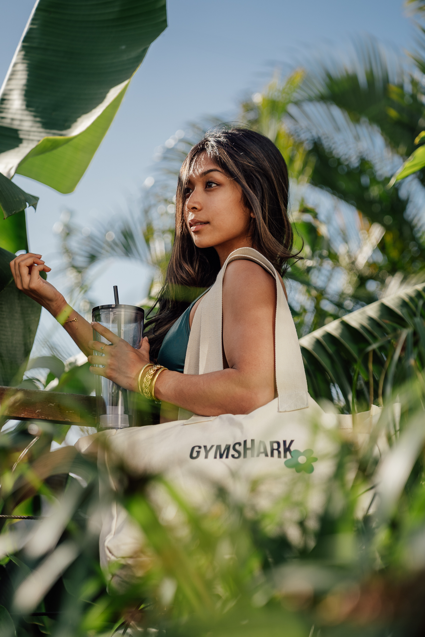 Naïla de profil entre des bananiers, tote bag Gymshark à l’épaule et gourde à la main, ciel bleu
