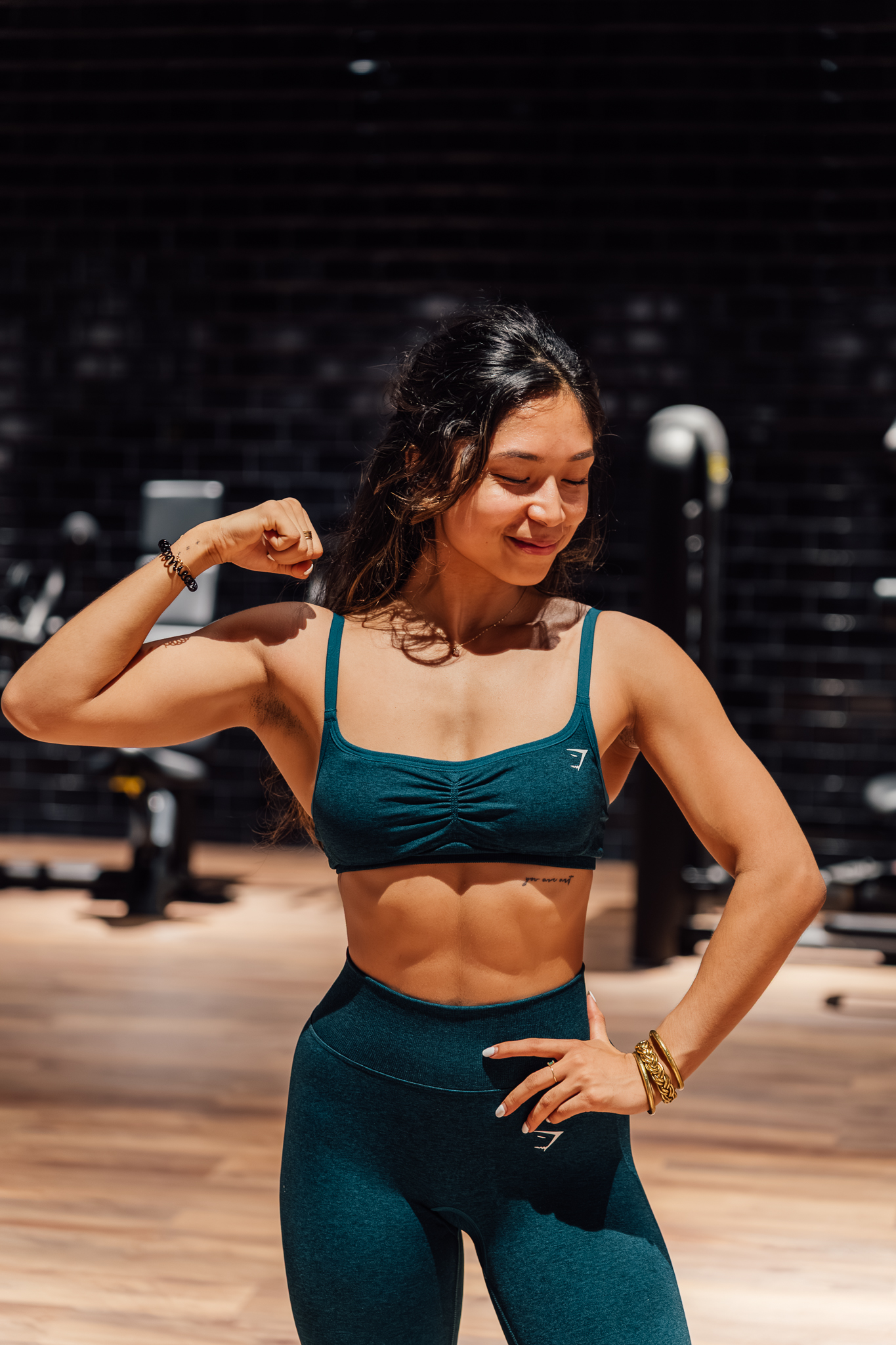 Naïla en tenue Gymshark bleu canard, souriante, le bras plié, dans une salle de sport aux lumières chaudes